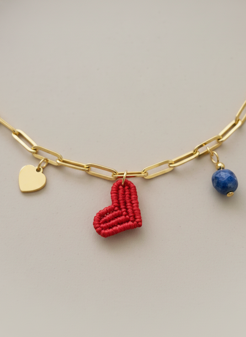Detalle del collar dorado con charm de corazón rojo tejido, colgante de corazón dorado y piedra natural azul en primer plano.