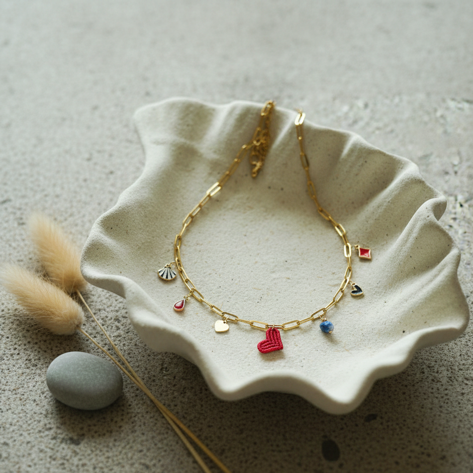 Collar dorado con charms variados en forma de corazón rojo tejido, concha, gota roja, piedra azul y abalorios esmaltados, sobre bandeja de resina en forma de estrella.