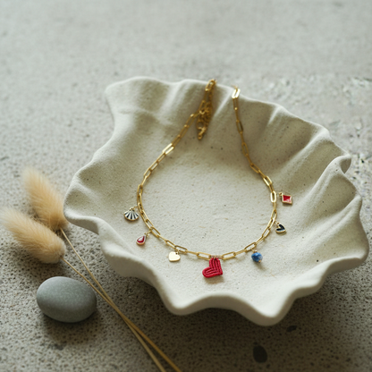 Collar dorado con charms variados en forma de corazón rojo tejido, concha, gota roja, piedra azul y abalorios esmaltados, sobre bandeja de resina en forma de estrella.