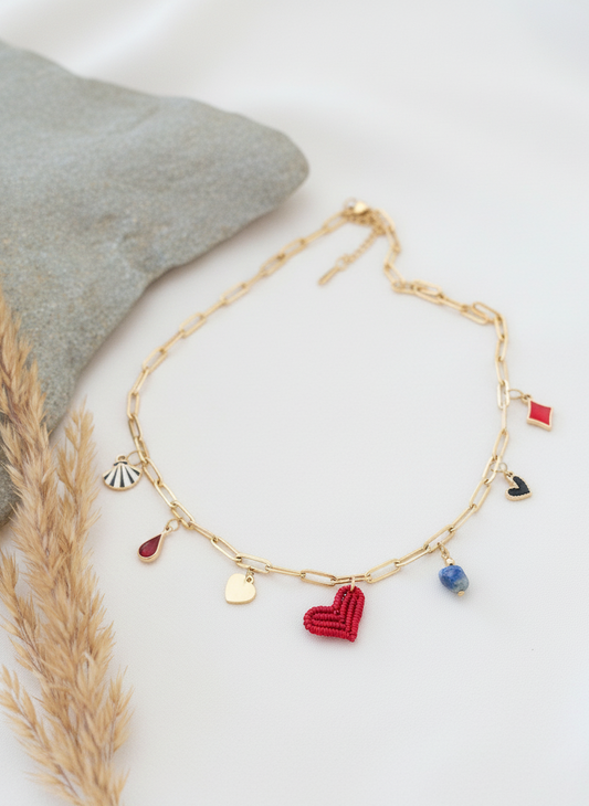 Collar dorado con charms variados en forma de corazón rojo tejido, concha, gota roja, piedra azul y abalorios esmaltados, sobre fondo neutro.