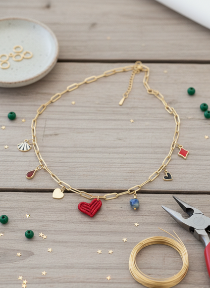 Collar dorado con charms variados en forma de corazón rojo de macrame, concha, gota roja, piedra azul y abalorios esmaltados, sobre fondo neutro.