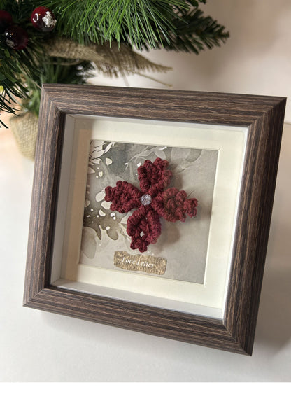 Mini cuadro cuadrado con flor de macramé burdeos. Muestra el detalle del fondo con textura de hoja o acuarela y el marco de caja oscura.