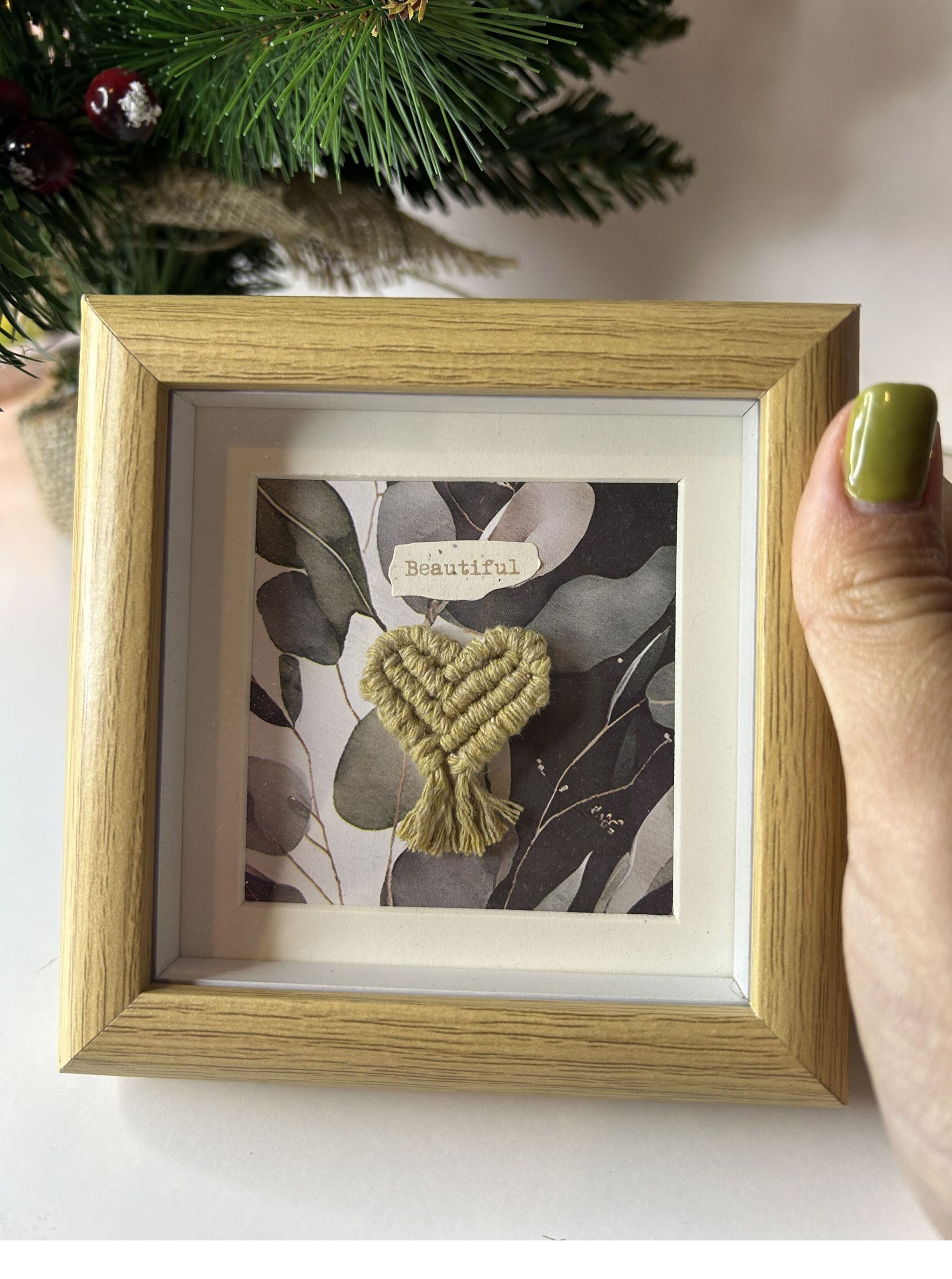 Persona sosteniendo el mini cuadro de macramé con corazón mostaza en el centro para mostrar su tamaño real. Detalle del arte en macramé y el diseño del marco de fotos.