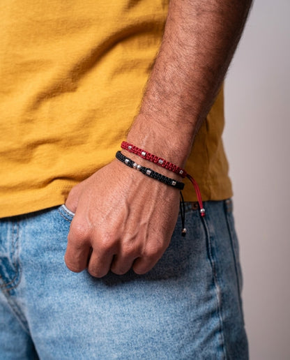 Pulseras dobles de macramé en rojo y negro con detalles metálicos, luciendo en muñeca masculina sobre camiseta mostaza