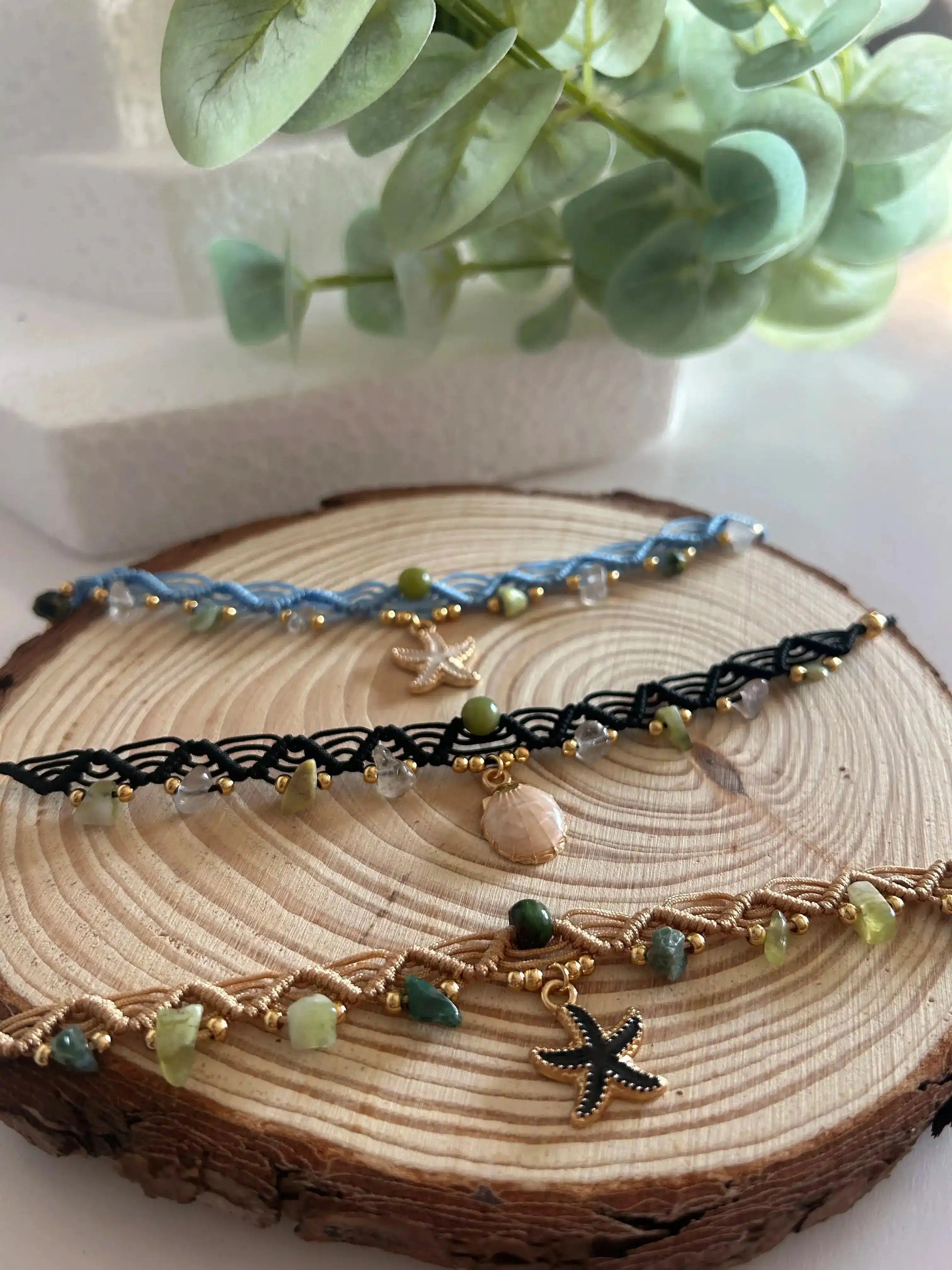 Chokers artesanales de macramé con hilo encerado, piedras naturales y dijes marinos sobre base de madera, decoración floral al fondo.