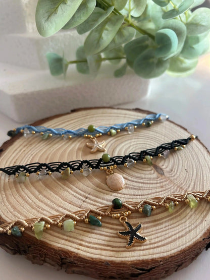 Chokers artesanales de macramé con hilo encerado, piedras naturales y dijes marinos sobre base de madera, decoración floral al fondo.