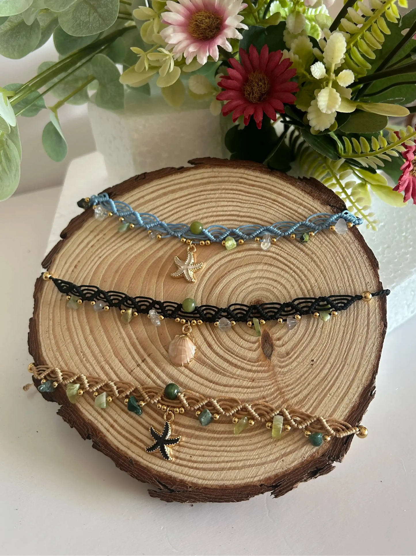 Chokers artesanales de macramé con hilo encerado, piedras naturales y dijes marinos sobre base de madera, decoración floral al fondo.