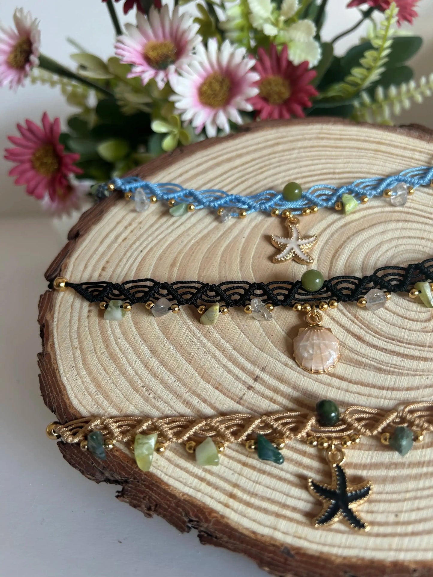 Chokers artesanales de macramé con hilo encerado, piedras naturales y dijes marinos sobre base de madera, decoración floral al fondo.
