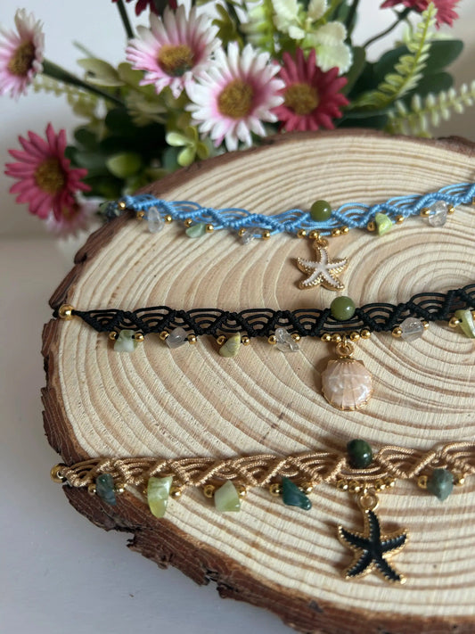 Chokers artesanales de macramé con hilo encerado, piedras naturales y dijes marinos sobre base de madera, decoración floral al fondo.